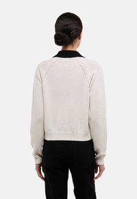 Pull couleur crème avec un col noir, présentant un design court, des poignets côtelés et des coutures raglan texturées. Le fond est uni.
