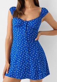 Abito estivo blu con fiori, maniche a sbuffo, scollatura a cuore e chiusura frontale con bottoni. Il tessuto presenta piccoli motivi floreali bianchi.