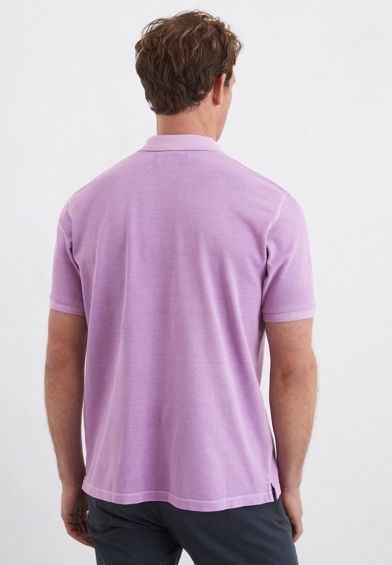 Liliowa koszulka polo z gładkiego materiału, z krótkimi rękawami i klasycznym kołnierzykiem. Zauważalne wcięcia po bokach na dolnym brzegu, zapewniające wentylację.