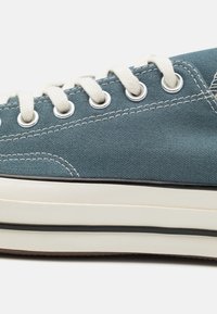 Blå lerretssneaker med hvite snørebånd, metalløyler, teksturert overflate, gummisåle og kontrasterende svart kant langs bunnen.