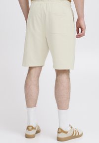 Light beige cotton shorts with an elastic waistband, featuring a back pocket. Worn with white socks and beige and brown sneakers.