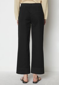 Černé kalhoty s širokými nohavicemi vyrobené z lehkého materiálu, s mírně vysokým pasem, zadními kapsami a čistými lemy. Nosí se s plochými černými sandály.