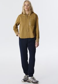 Mosterdkleurige hoodie-jack met een voorzak, gecombineerd met marineblauwe tapered broek en gestreepte sneakers. Het jack heeft een gladde afwerking en een trekkoord.