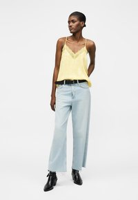 Γυναίκα που φοράει κίτρινη μπλούζα με δαντέλα, ανοιχτό μπλε τζιν παντελόνι με φαρδιά πόδια και ξεφτισμένες άκρες, μαύρη ζώνη και μαύρα μποτάκια αστραγάλου, στέκεται με το χέρι πίσω από την πλάτη.