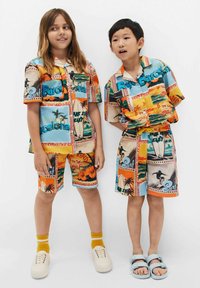Atitikmens vaikiškais drabužiais, kuriuose yra daugiaplanis, raštuotas audinys su tropiniais vaizdais, vandens sporto temomis ir tekstu, trumpomis rankovėmis marškiniai ir šortai.