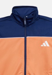 Σκούρο μπλε ναυτικό και πορτοκαλί αθλητικό μπουφάν με φερμουάρ, ψηλό γιακά και λευκό λογότυπο Adidas στο πορτοκαλί τμήμα του στήθους.
