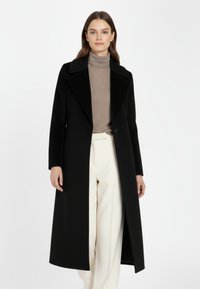 Vrouw gekleed in een lange zwarte trenchcoat over een taupe coltrui en witte high-waisted broek, staande tegen een effen witte achtergrond.