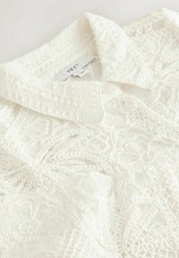 Chemise blanche en dentelle avec motif brodé floral, col boutonné et étiquette de marque "NEXT" visible à l’intérieur de l’encolure.
