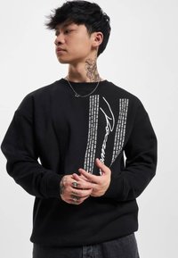 Black sweatshirt featuring a graphic design with white text and accents. The material appears soft and textured, with ribbed cuffs.