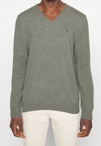 V-ringad tröja i olivgrön ullblandning, med långa ärmar, ribbad nederkant och en diskret logotyp på bröstet. Mjuk textur och åtsittande design.