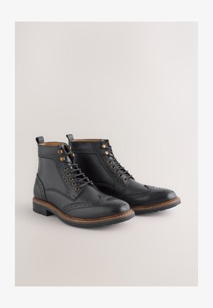 Botines negros de cuero con detalles brogue, puntera redonda, cierre de cordones al frente y suela marrón a contraste, con presillas para facilitar el calzado.