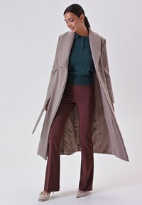 Cappotto lungo color beige chiaro con colletto a scollo, indossato sopra un polo verde scuro e pantaloni bordeaux. Il cappotto ha una texture liscia e un design elegante.