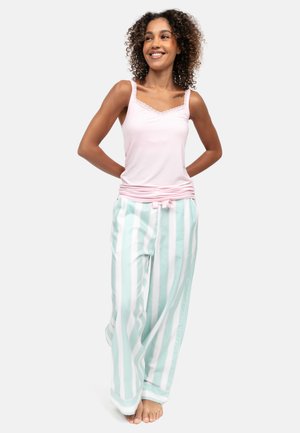 Lachende vrouw die blootsvoets staat, met krullend haar, een lichtroze mouwloze top en muntgroene en witte gestreepte pyjamabroek draagt.