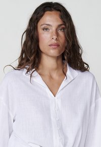 Camicia bianca di lino dal taglio rilassato, con chiusura a bottoni, colletto leggermente oversize e tessuto testurizzato.