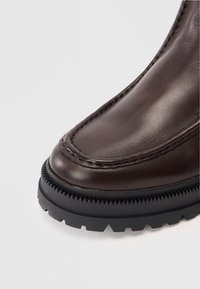 Bruine leren laars met een ronde neus, gedetailleerde stiksels en een getextureerde zwarte rubberen zool met een geribbeld patroon.