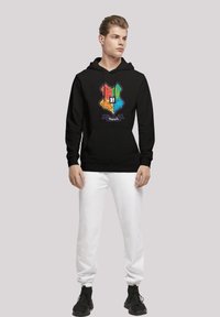 Mustra musta kapuutsiga, millel on mitmevärviline Hogwarts'i vapp, millel on kesksel "H" sümbol, koos valgete spordipükste ja mustade tossudega.