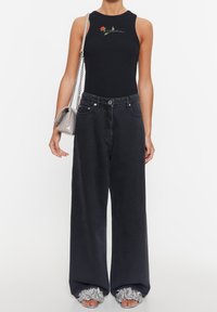 Nainen, joka pukeutunut mustaan hihattomaan yläosaan, jossa on kukkakuvio, leveälahkeisiin mustiin farkkuihin, hopeisiin röyhelösandaaleihin ja harmaaseen olkalaukkuun.