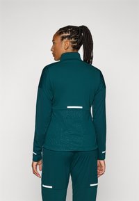 Veste de sport teal avec un col haut, des manches longues, un tissu texturé et un design abstrait au dos, avec des accents réfléchissants blancs.