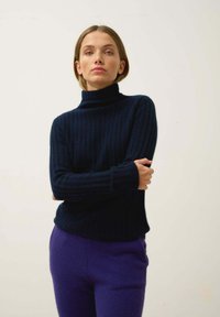 Granatowy sweter z golfem o prążkowanej fakturze, z długimi rękawami, zestawiony z fioletowymi spodniami o wysokim stanie. Miękka tekstura, dopasowany krój.