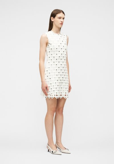 Γυναίκα όρθια, φορώντας αμάνικο λευκό δαντελένιο φόρεμα με floral μοτίβο και λευκά παπούτσια με χαμηλό τακούνι και αγκράφες, μπροστά σε απλό φόντο.