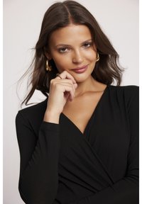 Czarna bluzka z długimi rękawami, z głębokim dekoltem w kształcie litery V i gładką teksturą. Złote kolczyki w kształcie kół i subtelna ręka podkreślają projekt.