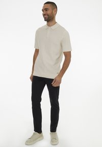 Polo s krátkým rukávem v texturované krémové tkanině s límečkem a tříknoflíkovou légou, doplněné o černé slim-fit kalhoty a světlé tenisky.