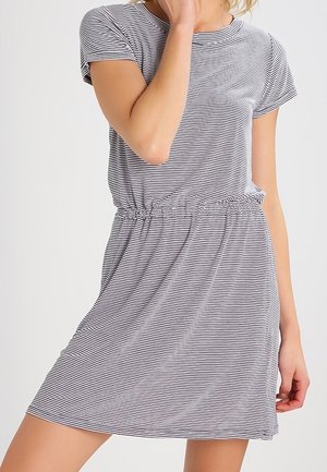 Vrouw met een zwart-wit horizontaal gestreepte korte mouw jurk met een elastische tailleband, staand tegen een effen achtergrond.