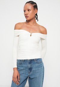 White off-the-shoulder top with long sleeves, featuring a central keyhole accent and gathered texture. Paired with blue denim jeans.