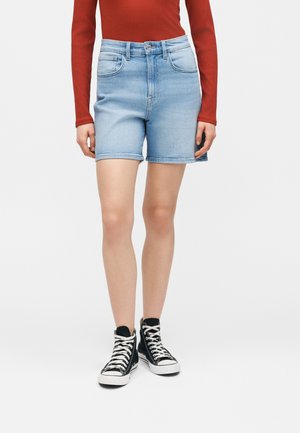 Person iført lyseblå denimshorts, en langærmet kongeblå top og sorte højt-siddende sneakers, stående mod en hvid baggrund.