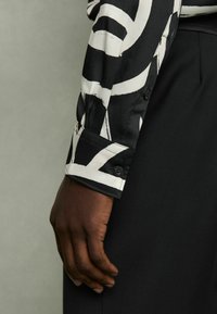 Manica di camicia a righe bianche e nere con polsino abbottonato; texture del tessuto liscia; design elegante e aderente; mano appoggiata su una superficie nera.
