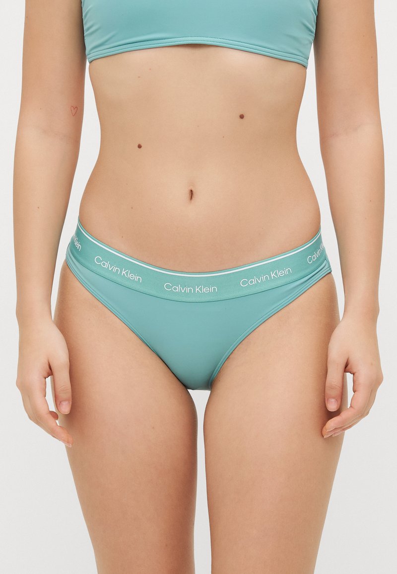 Teal Calvin Klein bikini-bukse med elastisk midje og hvit logo-tekst, glatt stoff og klassisk snittdesign.
