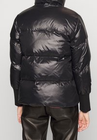 Doudoune noire avec une texture brillante, dotée de compartiments cousus, d'une capuche et de poignets côtelés. Associée à un pantalon sombre et brillant.