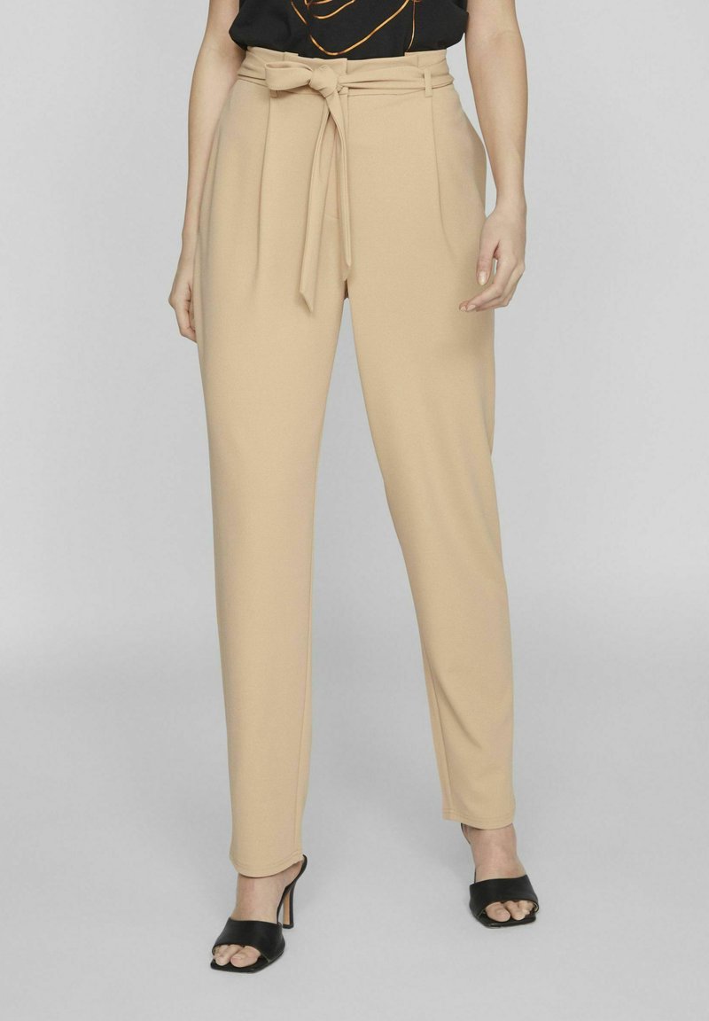 Vrouw gekleed in beige high-waisted broek met een gestrikt ceintuur en zwarte open teenhakken, staand tegen een lichtgrijze achtergrond.