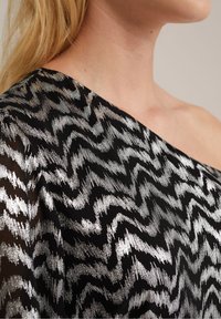 Černý látkový top se stříbrným cikcak vzorem, s asymetrickým výstřihem a průhlednou texturou. Důraz na dynamický design a kontrastní barvy.