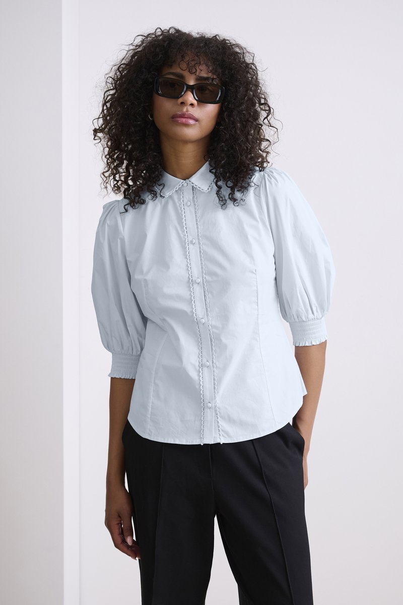 Blusa azzurro chiaro con maniche a palloncino, colletto, chiusura frontale con bottoni, bordi decorativi e polsini arricciati, indossata con pantaloni neri.