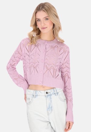 Nő világos lila, gombos, rövidített kötött kardigánban és világos, magas derekú, koptatott farmerben, egyszínű háttér előtt.