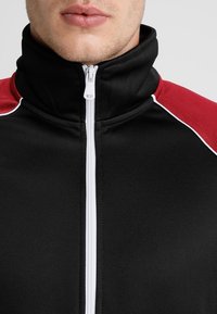 Giacca nera con zip e colletto alto; presenta accenti rossi a contrasto sulle spalle e piping bianco lungo le cuciture. Tessuto flessibile.