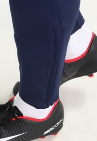 Botín de fútbol negro con acentos rojos, combinado con calcetines blancos y pantalones deportivos azul marino con detalle de abertura lateral.