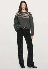 Jeune femme portant un pull en maille gris à motif, un pantalon en velours noir avec une ceinture, et des mocassins noirs, debout devant un fond uni.
