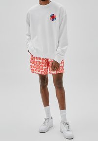 Witte sweatshirt met een multicolor logo op de borst, gecombineerd met rode shorts met een herhalend wit grafisch patroon, gedragen met witte sneakers.