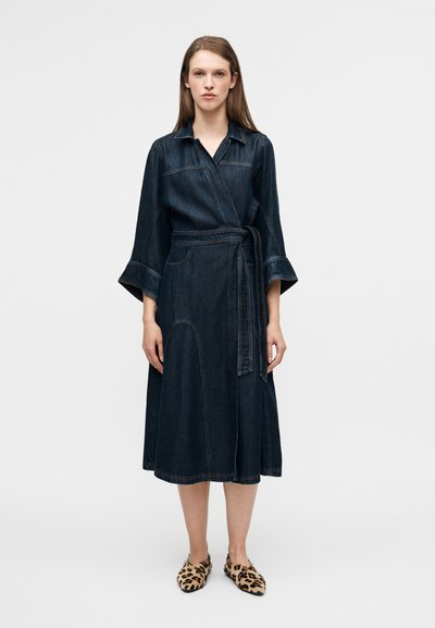 Robe portefeuille en jean avec de longues manches évasées, teinte bleu foncé, ceinture nouée à la taille, et surpiqûres contrastantes. Associée à des chaussures à imprimé léopard.