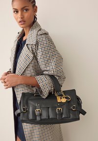 Melns ādas rokassomas ar zelta aksesuāriem, tai ir trīs priekšējās kabatas, noņemams siksniņa un dekoratīvs atslēgu piekariņš.