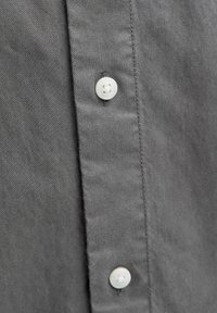 Pelēks krekls ar pogām, kam ir diagonālas auduma tekstūras, ar baltām pogām un tuvplānu uz pogu sloksni un šuvju detaļām.