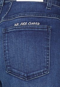 Vaqueros de mezclilla azul oscuro con un bolsillo trasero que presenta un texto bordado en blanco que dice "ESTAMOS CERRADOS." Corte clásico con costuras detalladas.