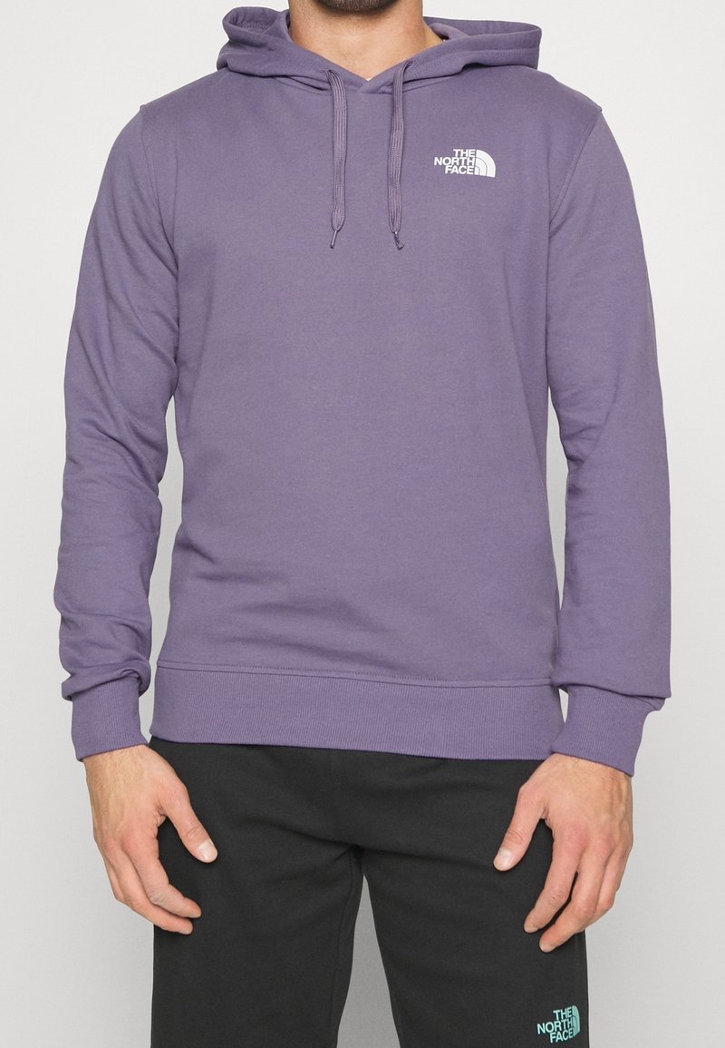 Homme portant un sweat à capuche violet et un pantalon noir, tous deux avec le logo blanc The North Face sur le côté gauche.