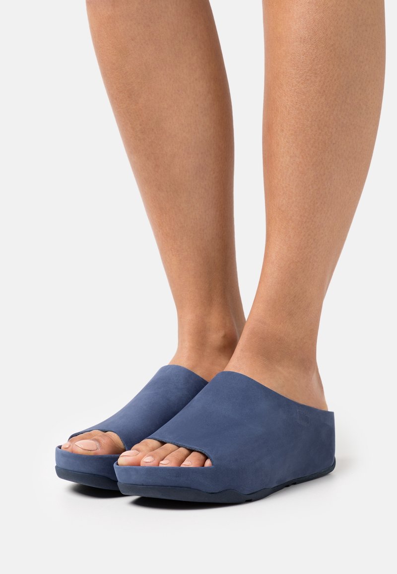 Jambes portant des sandales bleues à enfiler, à bout ouvert avec des semelles épaisses sur un fond uni clair.