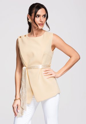 Vrouw met donker haar draagt een mouwloos beige topje met asymmetrische zoom en satijnen tailleband, gecombineerd met een witte broek, poserend tegen een effen achtergrond.