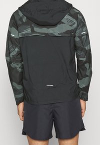 Zwarte lichte jacket met een camouflagepatroon op de mouwen, voorzien van een capuchon, voorzak en een opvouwbaar ontwerp.