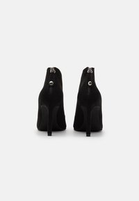 Botines de ante negros con tacón alto, que cuentan con una cremallera lateral y un acento metálico en el tacón. Textura suave y silueta estilizada.