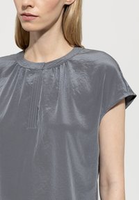 Graues Kurzarmblusenshirt mit rundem Ausschnitt, ausgestattet mit Schulterfalten und subtilen Nähdetails. Hergestellt aus glänzendem, glattem Stoff.